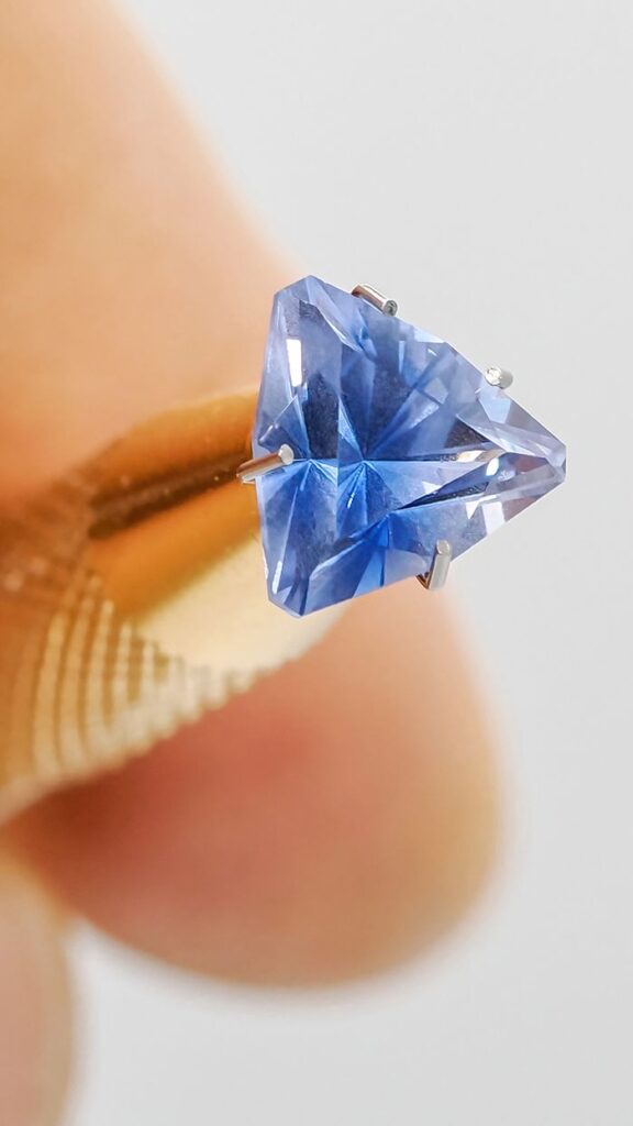 Trilliant cut blue sapphire held by tweezers, showcasing precision and color.